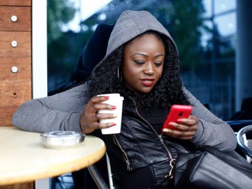 Woman seated outdoors at a round table, using a red cell phone in her left hand and holding a coffee in her right hand.