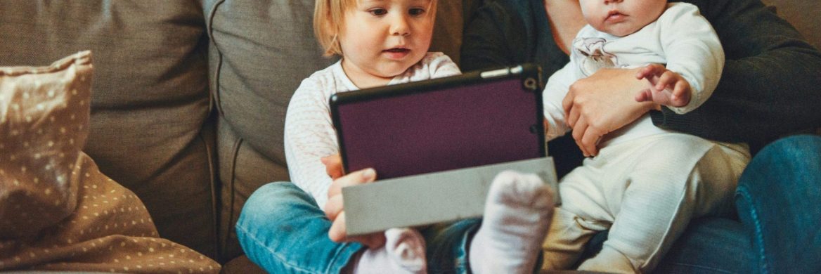 A woman looks at a tablet while she holds two children.