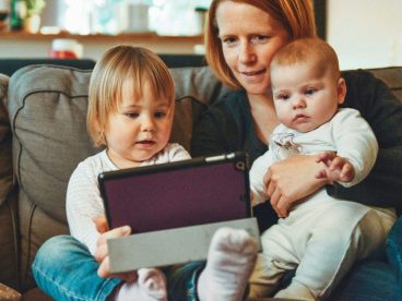 A woman looks at a tablet while she holds two children.