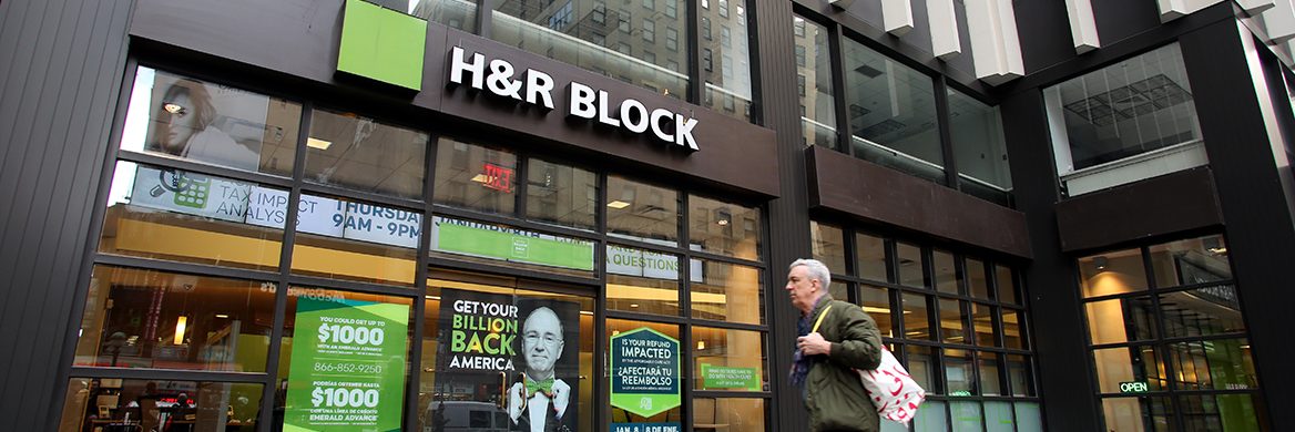 “A man walking in front of an H&R Block storefront with multiple ads promoting paid tax preparation.