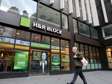 “A man walking in front of an H&R Block storefront with multiple ads promoting paid tax preparation.
