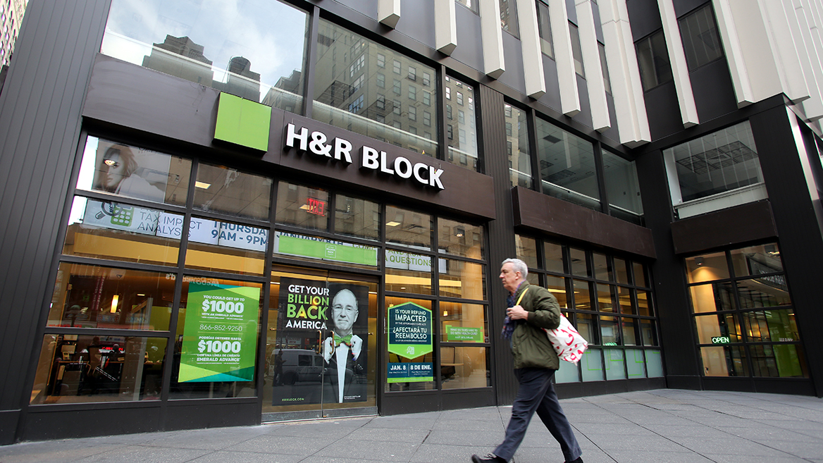 “A man walking in front of an H&R Block storefront with multiple ads promoting paid tax preparation.