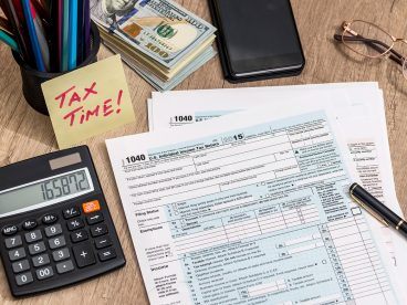 A 1040 tax return document is on a desk next to a calculator, a stack of money, a smartphone, and a sticky note that reads, “Tax Time!”.