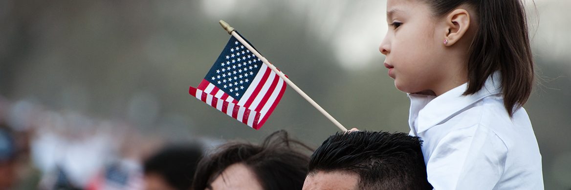 Young daughter sitting on her father’s shoulders waiving an American Flag.