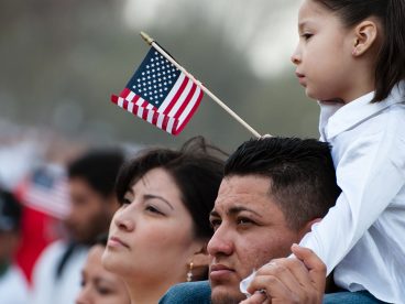 Young daughter sitting on her father’s shoulders waiving an American Flag.