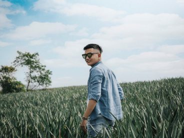 Person with sunglasses on standing in a field looking over their shoulder toward the camera.