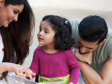 A mother talking to her daughter while her father listens, kneeling behind the child.