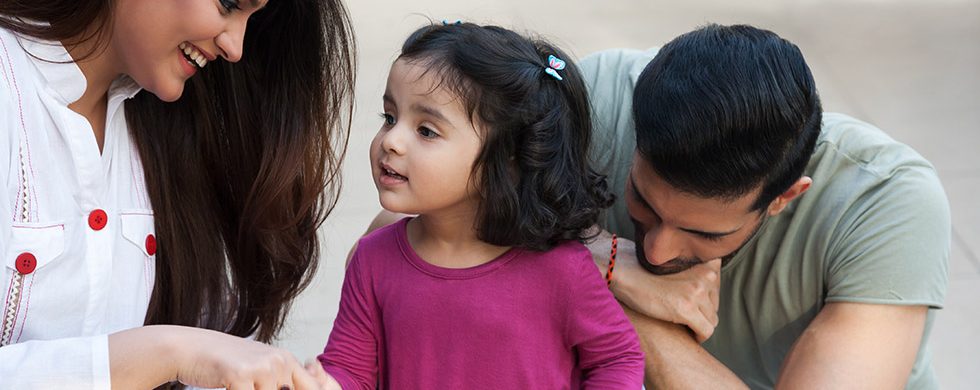 A mother talking to her daughter while her father listens, kneeling behind the child.