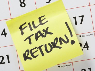 Yellow post-it note with “FILE TAX RETURN!” written in marker placed on a calendar in between the 14th and 17th days.