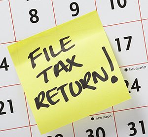 Yellow post-it note with “FILE TAX RETURN!” written in marker placed on a calendar in between the 14th and 17th days.